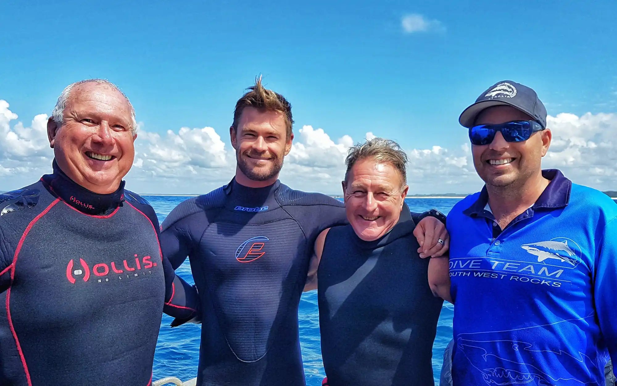 Four people wearing wetsuits and water gear standing together against an ocean backdrop.